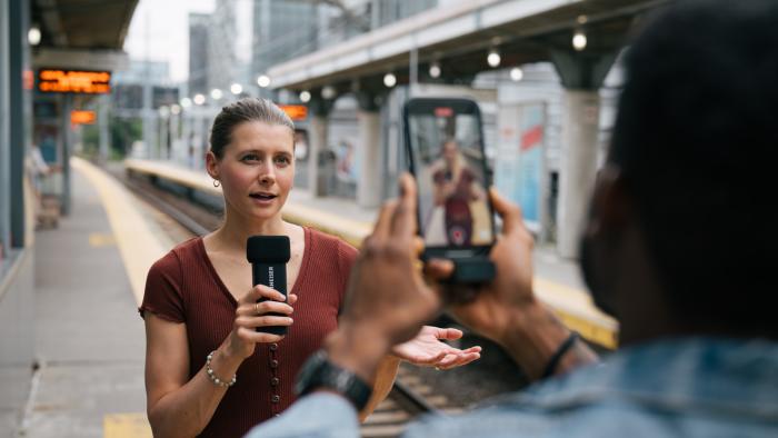 A person holding a mic, with another person filming them on a phone.