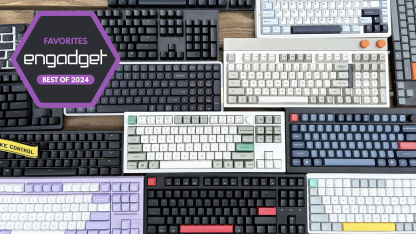 A wide selection of mechanical keyboards in different sizes, shapes and colors sit in close proximity to one another on top of a brown wooden table.