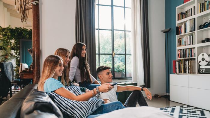 Four friends watching tv at home. They are sitting on the sofa, taking a break from homework.