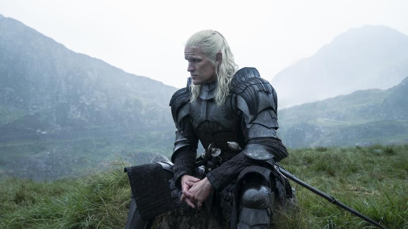 Still from House of the Dragon. A man with long white hair wears a suit of armor as he sits on a mound of grass. There are mountains in the background.