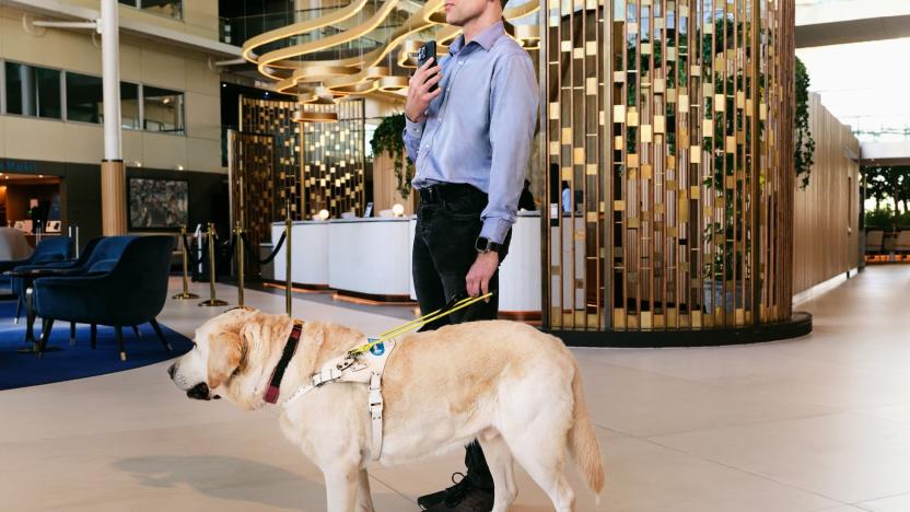 A man holds a smartphone to his chest with the rear camera facing outward as he stands with a guide dog in a hotel lobby.