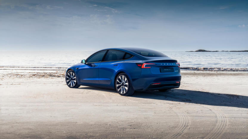 A blue Tesla sedan on a beach. Tire tracks in the sand.