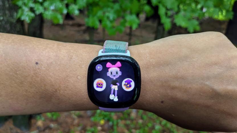 The Fitbit Ace LTE pictured on a wrist. Its display shows an "eejie," or virtual companion. The Eejie is a bald humanoid with purple skin, large eyes and fangs and wears a large pink bow on its head, black T shirt, light orange and white shorts, and white bunny slippers