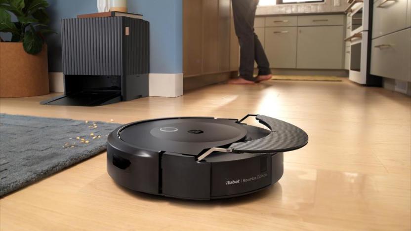 Product lifestyle photo of the Roomba Combo 10 Max + Autowash Dock. The robot vacuum glides across the floor as a person works in the kitchen behind.