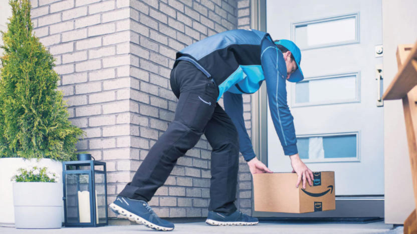 A person placing an Amazon-branded box on a welcome mat in front of a white door.