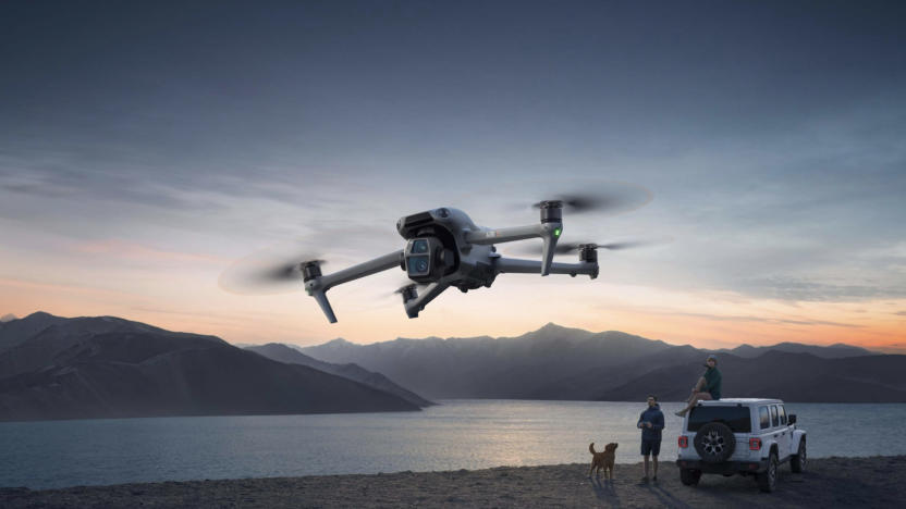 A small flying machine against the sky with two humans, a dog and a vehicle in the background.