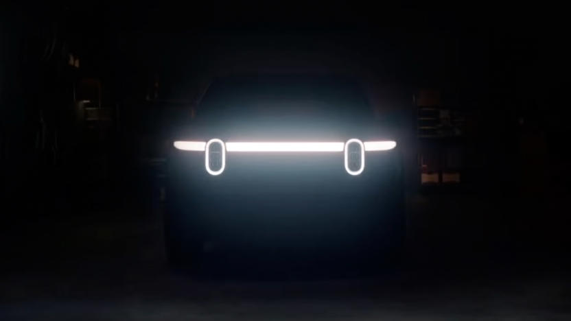 Teaser image for the Rivian R2 EV. Two vertically oval headlights along a sleek horizontal light bar hint at the front of the vehicle. Otherwise nearly black image of a garage.