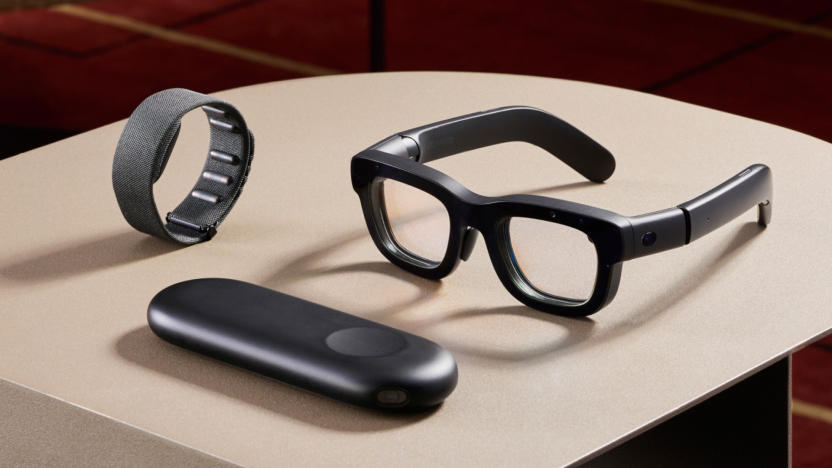 A pair of Meta's Orion AR glasses on a table, with two accessories also on display. A wrist-strap with a module on its underside sits to the left of the glasses while a black, rounded rectangular device with an indented circle sits in front.