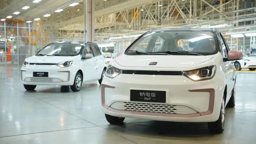 Large car factory or showroom floor featuring two small white electric vehicles. Scaffolding and other cars visible in the background.