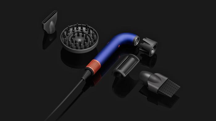 The components of the Dyson Supersonic R hairdryer are laid out next to each other on a black surface. in the middle is the main J-shaped tubular dryer body with its several attachments.